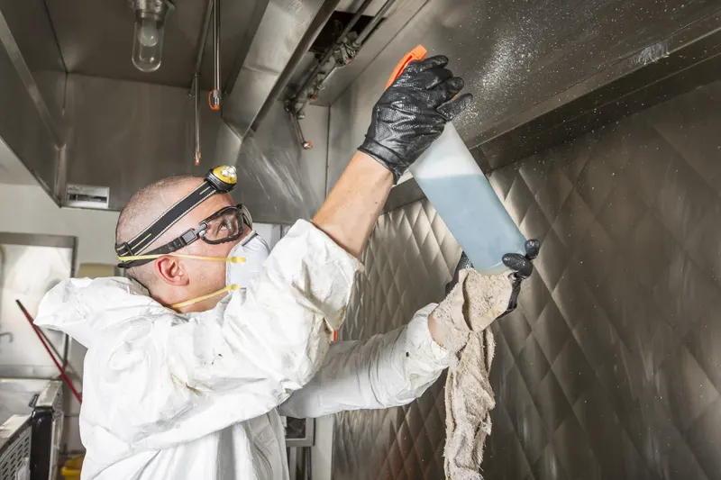 Technician spraying overhead inside a commercial exhaust hood with a cleaning cloth in hand