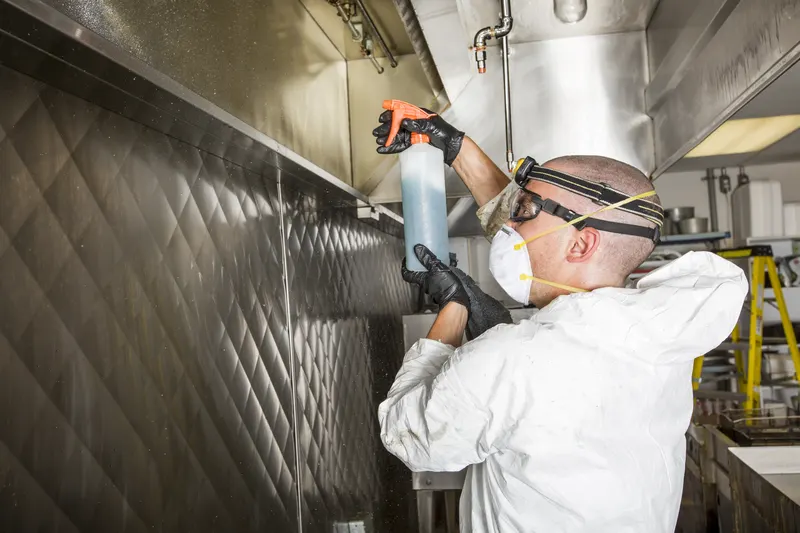 Technician in coveralls spraying degreaser onto the inside face of a stainless steel exhaust canopy
